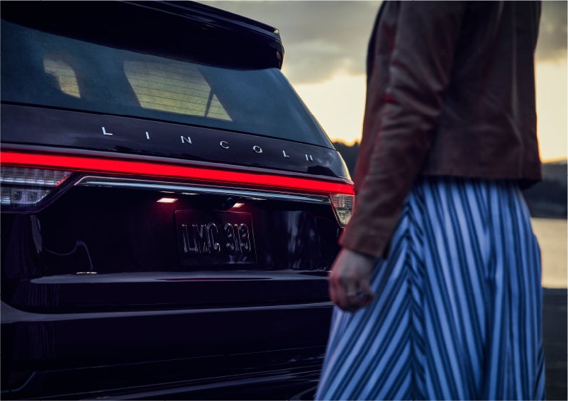 A person is shown near the rear of a 2023 Lincoln Aviator® SUV as the Lincoln Embrace illuminates the rear lights | Lincoln of New Bern in New Bern NC